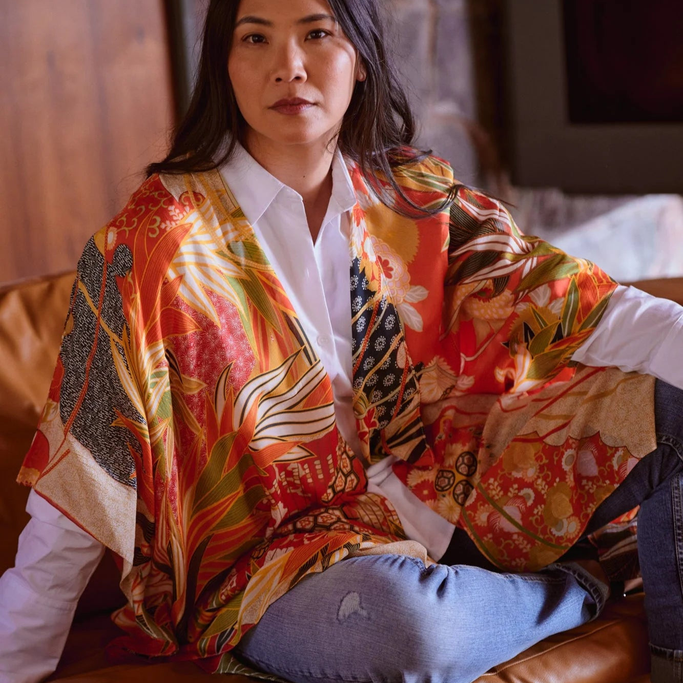 Woman wearing a colorful patterned shawl over a white shirt and jeans, sitting on a brown leather couch.
