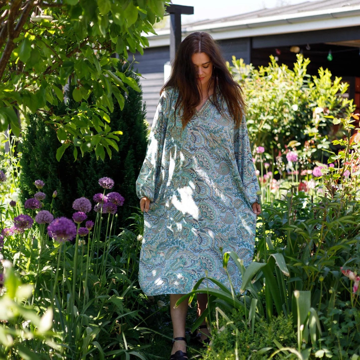 Woman in a light blue dress walking through a garden with flowers and trees.