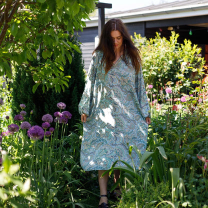 Woman in a light blue dress walking through a garden with flowers and trees.