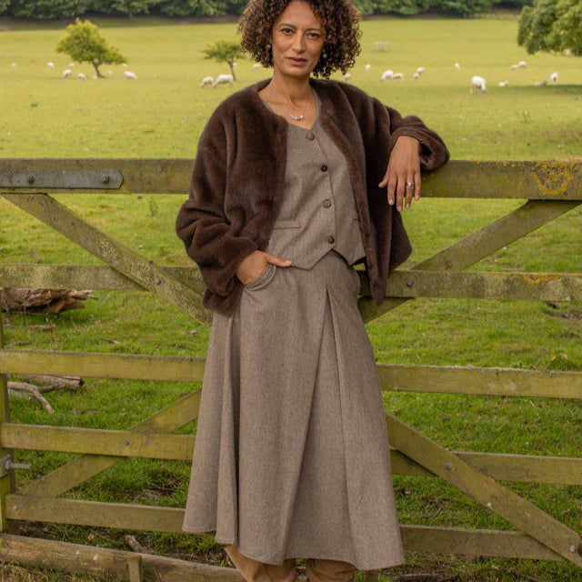 Woman in a brown coat and gray dress standing in a field with sheep and greenery.